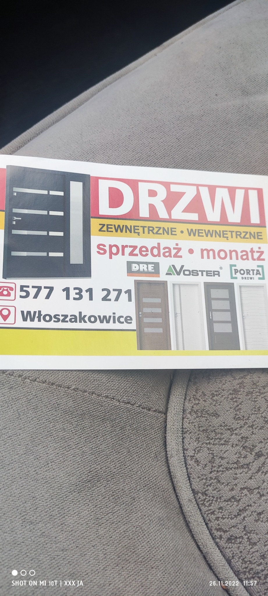 Wizytówka firmy oferującej sprzedaż i montaż drzwi zewnętrznych i wewnętrznych, z numerem telefonu, adresem i logotypami producentów DRE, VOSTER i PORTA, leżąca na szarej tapicerce.