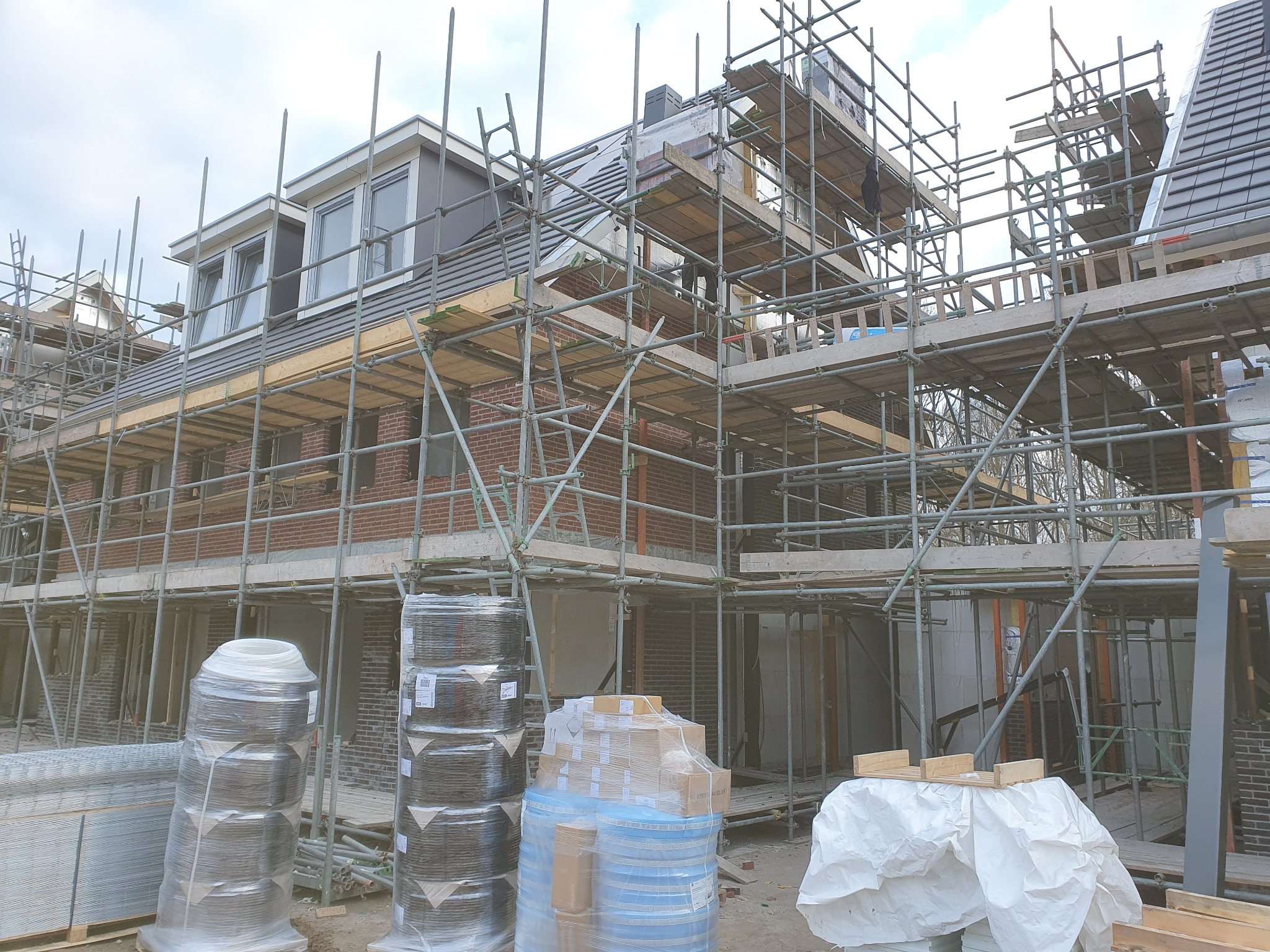 Budowa domu jednorodzinnego w trakcie murowania ścian, widoczne rusztowania, cegły, materiały budowlane zabezpieczone folią, pochmurne niebo.