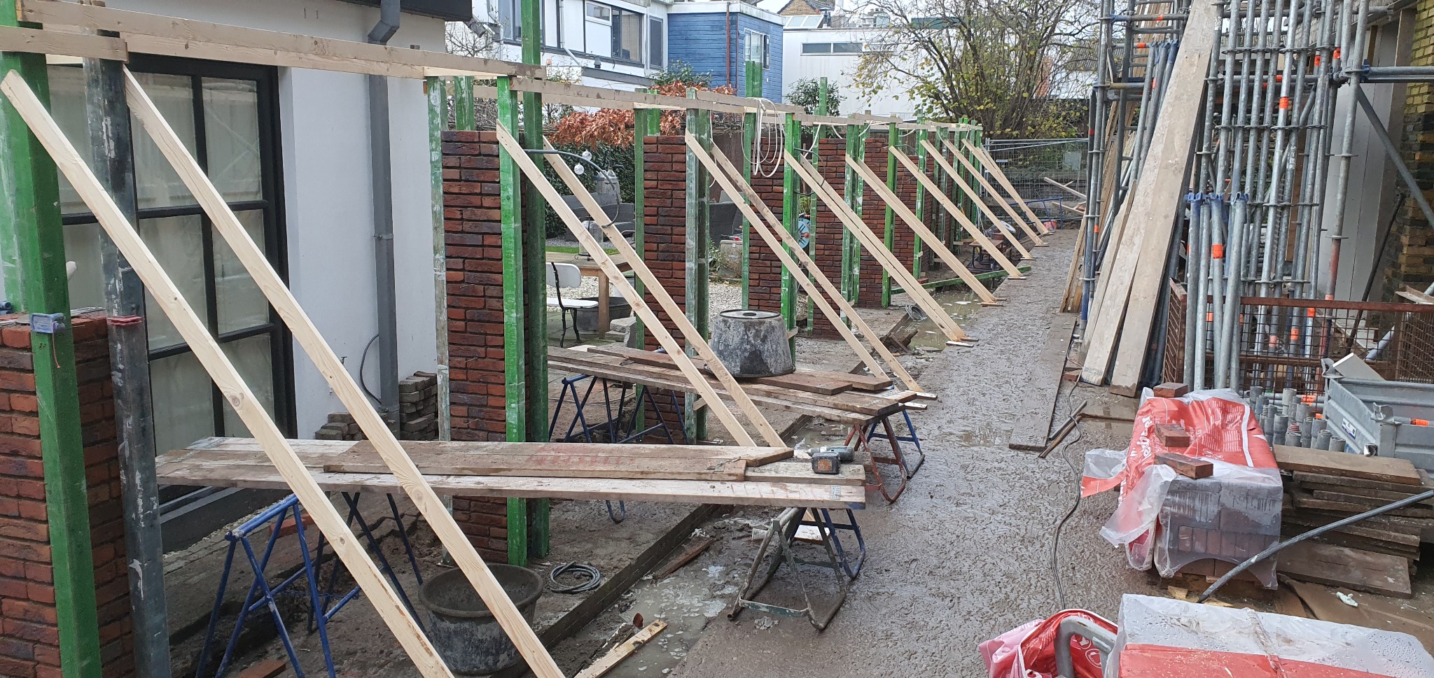 Wznoszone z czerwonej cegły filary ścian z drewnianym szalunkiem, widoczne rusztowania i materiały budowlane na mokrym podłożu.