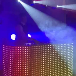 Osoba obsługująca konsoletę DJ-ską podświetloną kolorowymi diodami, oświetloną reflektorami i wypełnioną dymem scenicznym.