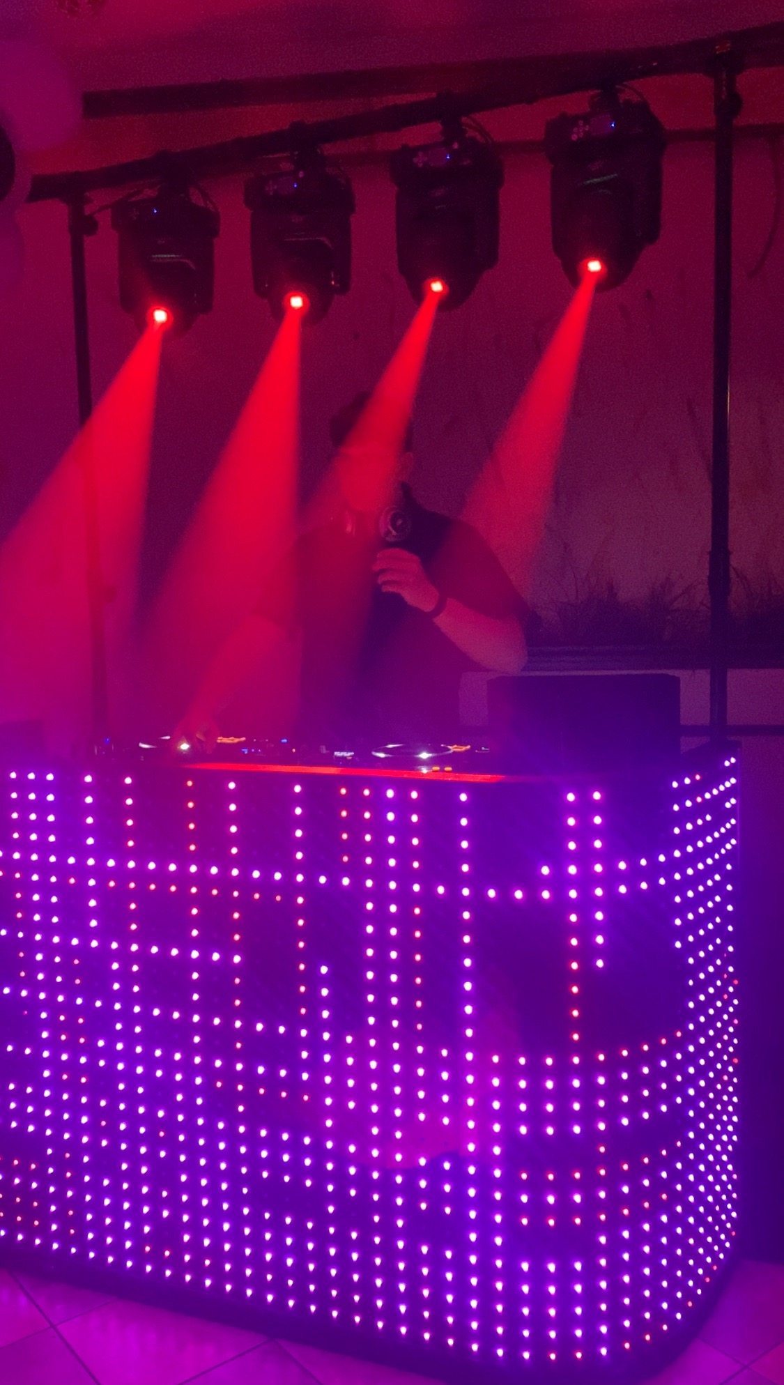 DJ za podświetlonym panelem LED, reflektory z czerwonym światłem skierowane w dół, tworzące atmosferę imprezy.