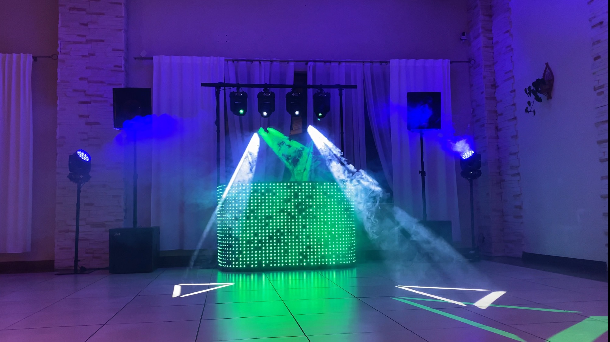Podświetlony ledami panel DJ-ski z wytwornicą dymu i reflektorami na scenie, w tle białe zasłony i ściana z cegły.