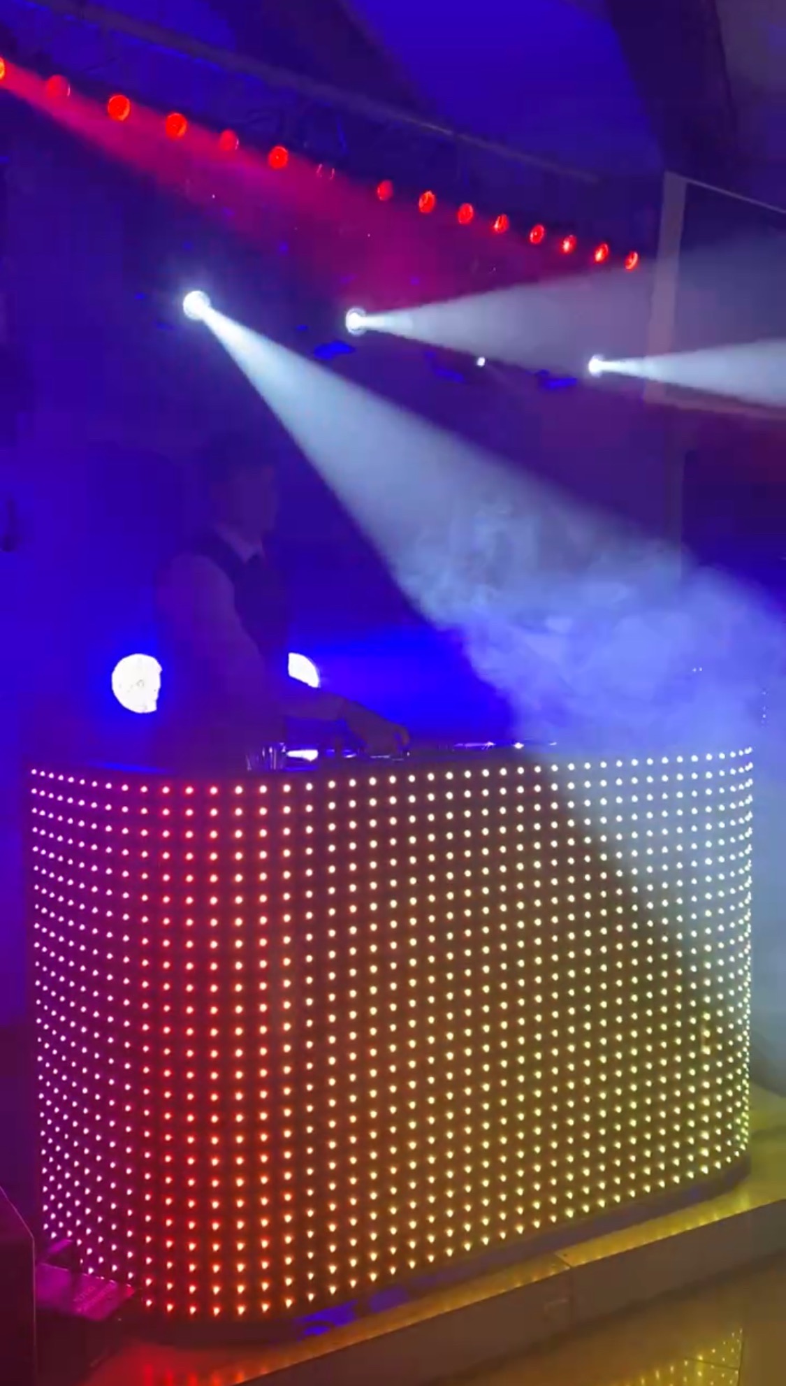 Osoba obsługująca konsoletę DJ-ską podświetloną kolorowymi diodami, oświetloną reflektorami i wypełnioną dymem scenicznym.