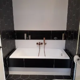 Nowoczesna łazienka z białą wanną obudowaną czarnymi panelami, mozaiką heksagonalną na ścianach i podłodze, oraz armaturą w kolorze różowego złota.