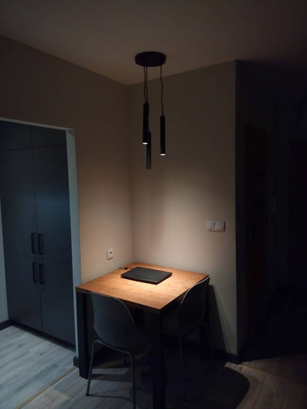 Wnętrze pokoju z drewnianym stołem, dwoma szarymi krzesłami, zamkniętym laptopem na blacie i trzema wiszącymi, czarnymi lampami tubami pod sufitem, obok zabudowana szafa.