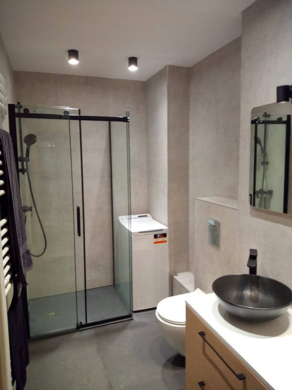 Nowoczesna łazienka z kabiną prysznicową typu walk-in, umywalką nablatową w kolorze czarnym, pralką ukrytą za kabiną oraz szarą, minimalistyczną kolorystyką ścian i podłogi.