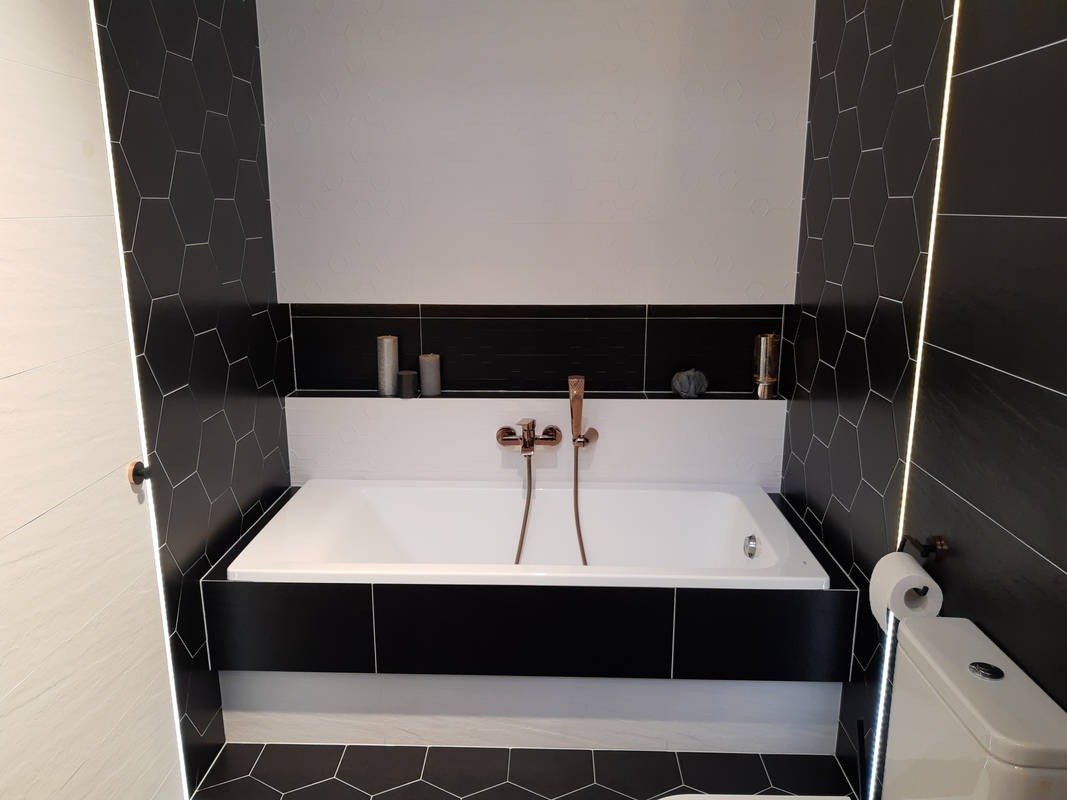 Nowoczesna łazienka z białą wanną obudowaną czarnymi panelami, mozaiką heksagonalną na ścianach i podłodze, oraz armaturą w kolorze różowego złota.