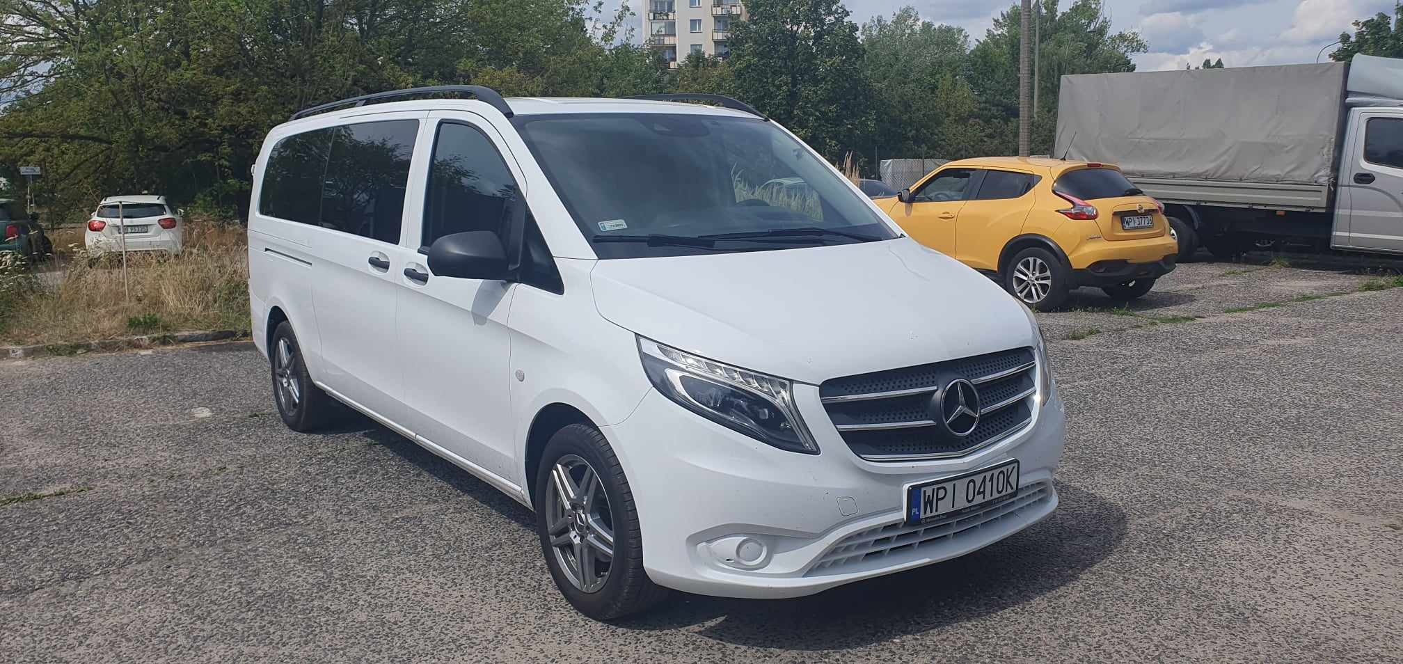 Biały Mercedes Vito z bagażnikiem dachowym, zaparkowany na parkingu, obok żółty Nissan Juke i szary samochód dostawczy z plandeką.