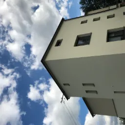 Nowoczesny budynek z jasną elewacją widziany z dołu, z wyraźnym podziałem na dwie barwy, na tle błękitnego nieba z białymi chmurami.