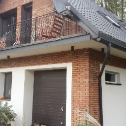 Dwukondygnacyjny dom z ceglaną elewacją, balkonem z kutą balustradą i garażem z brązową bramą segmentową.