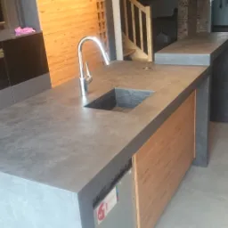 Szary blat wyspy kuchennej z wbudowanym zlewem i chromowaną baterią, obudowany drewnopodobnymi panelami, na tle schodów i ceglanej ściany, widoczny fragment zabudowy kuchennej z ekspresem do kawy.