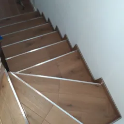 Schody wyłożone brązowymi płytkami imitującymi drewno, z metalowymi listwami na krawędziach stopni, widziane z góry pod kątem.