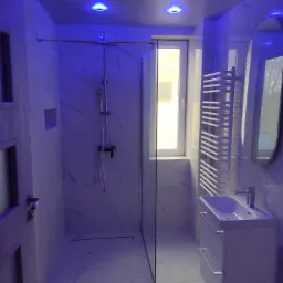 Nowoczesna łazienka z kabiną prysznicową typu walk-in, umywalką z szafką i grzejnikiem drabinkowym, oświetlona niebieskim światłem LED.