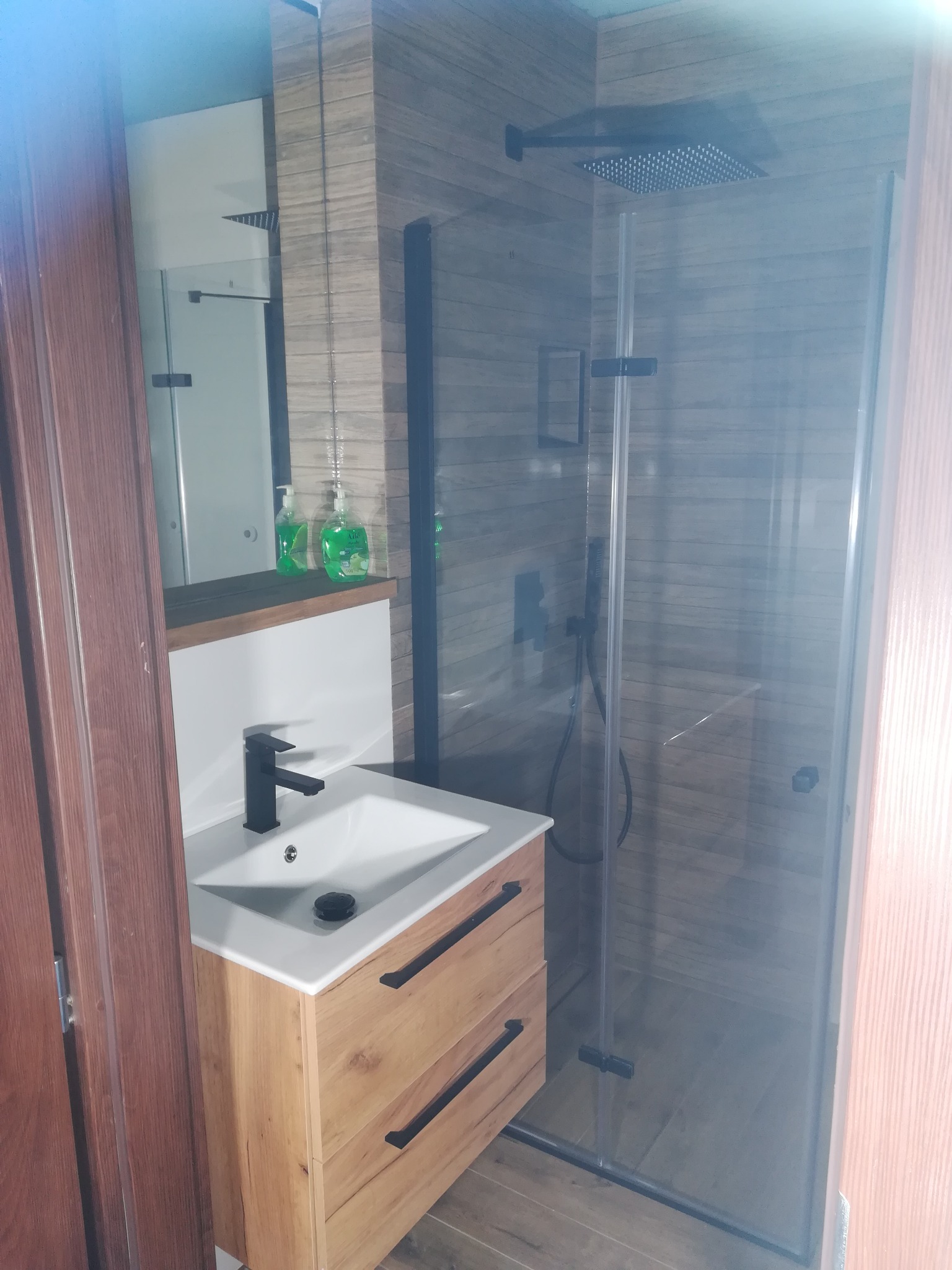 Nowoczesna łazienka z kabiną prysznicową typu walk-in, drewnopodobne płytki na ścianach i podłodze, umywalka nablatowa z szafką z drewnianym fornirem i czarną armaturą.