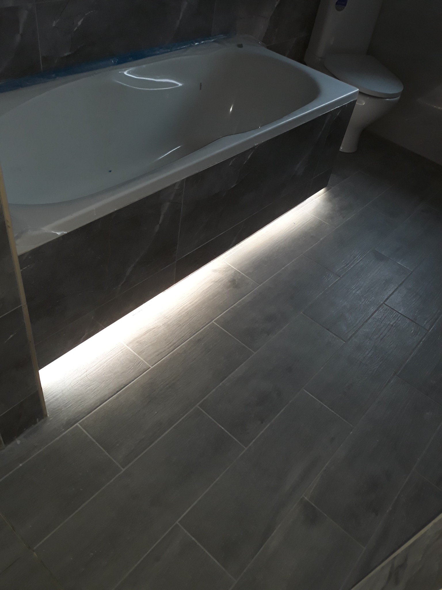 Nowoczesna łazienka z podświetloną wanną i ciemną, drewnopodobną glazurą na podłodze i obudowie wanny, widoczna również toaleta.