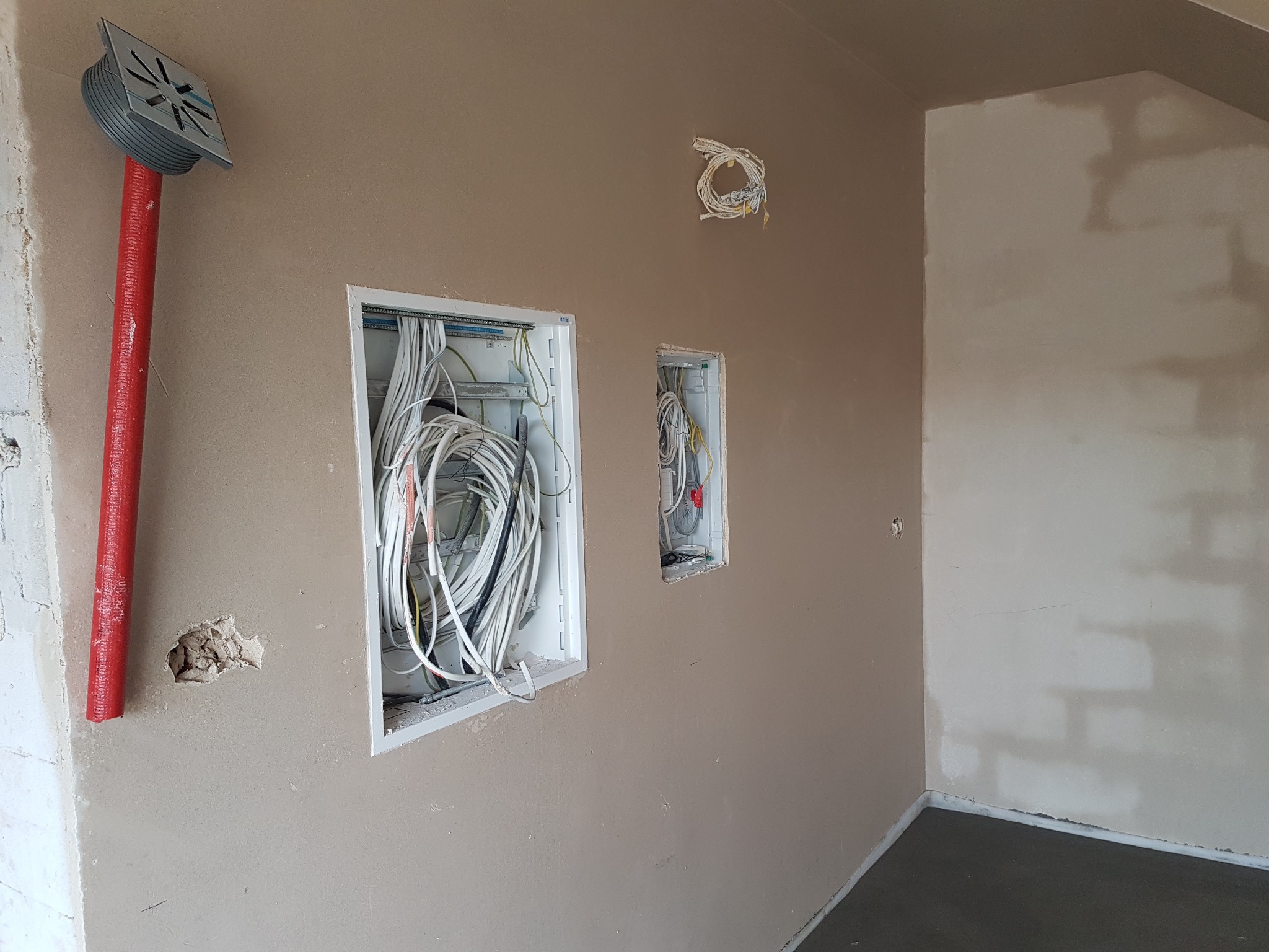 Ściana wewnętrzna w trakcie prac wykończeniowych: świeżo położony tynk, widoczne puszki elektryczne z okablowaniem oraz pionowa rura kanalizacyjna z metalową kratką w górnej części.