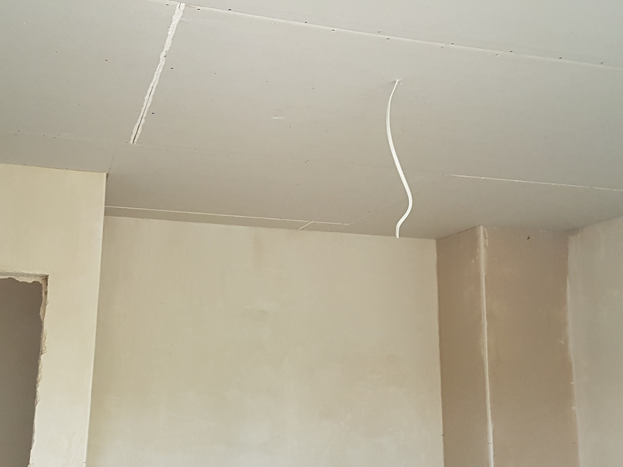 Wnętrze w trakcie wykończenia: sufit z płyt kartonowo-gipsowych z widocznymi łączeniami, wystający kabel elektryczny i fragment ściany w jasnym odcieniu beżu z surowym otworem drzwiowym.