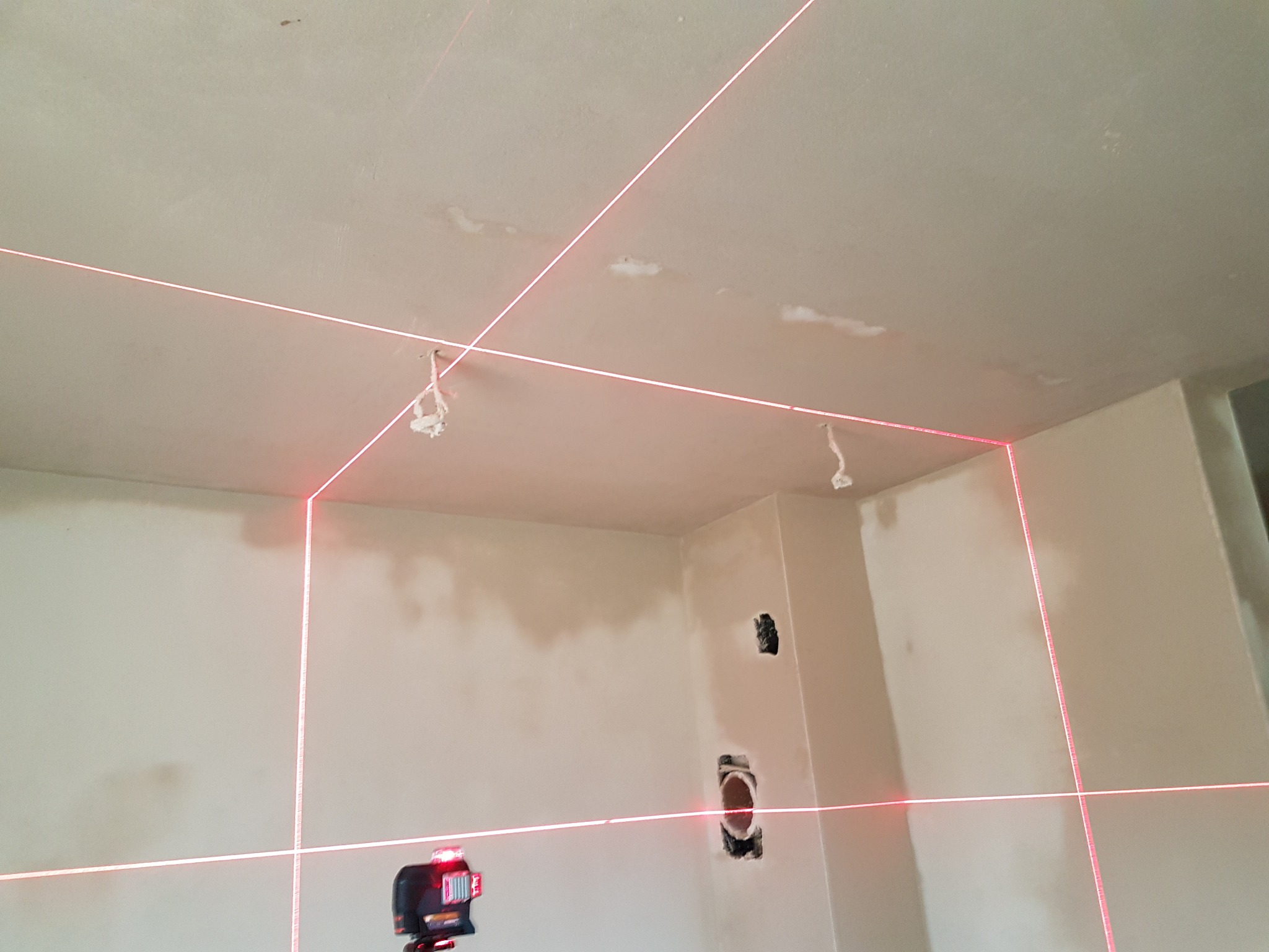 Wnętrze w trakcie remontu z widocznymi laserowymi liniami poziomymi i pionowymi, wyznaczającymi płaszczyzny na ścianach i suficie, oraz otworami pod instalacje elektryczne.