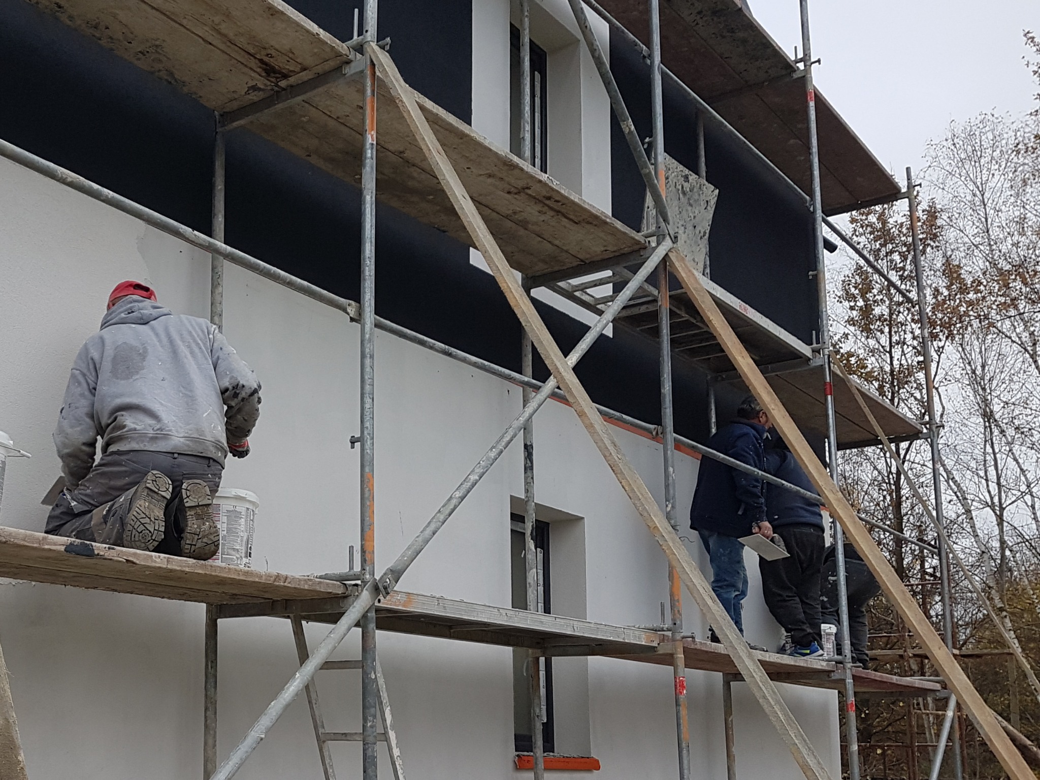 Pracownicy na rusztowaniu tynkują elewację domu jednorodzinnego, widoczne ocieplenie w czarnym kolorze, jeden z pracowników nakłada tynk pacą, obok stoją wiadra z zaprawą.