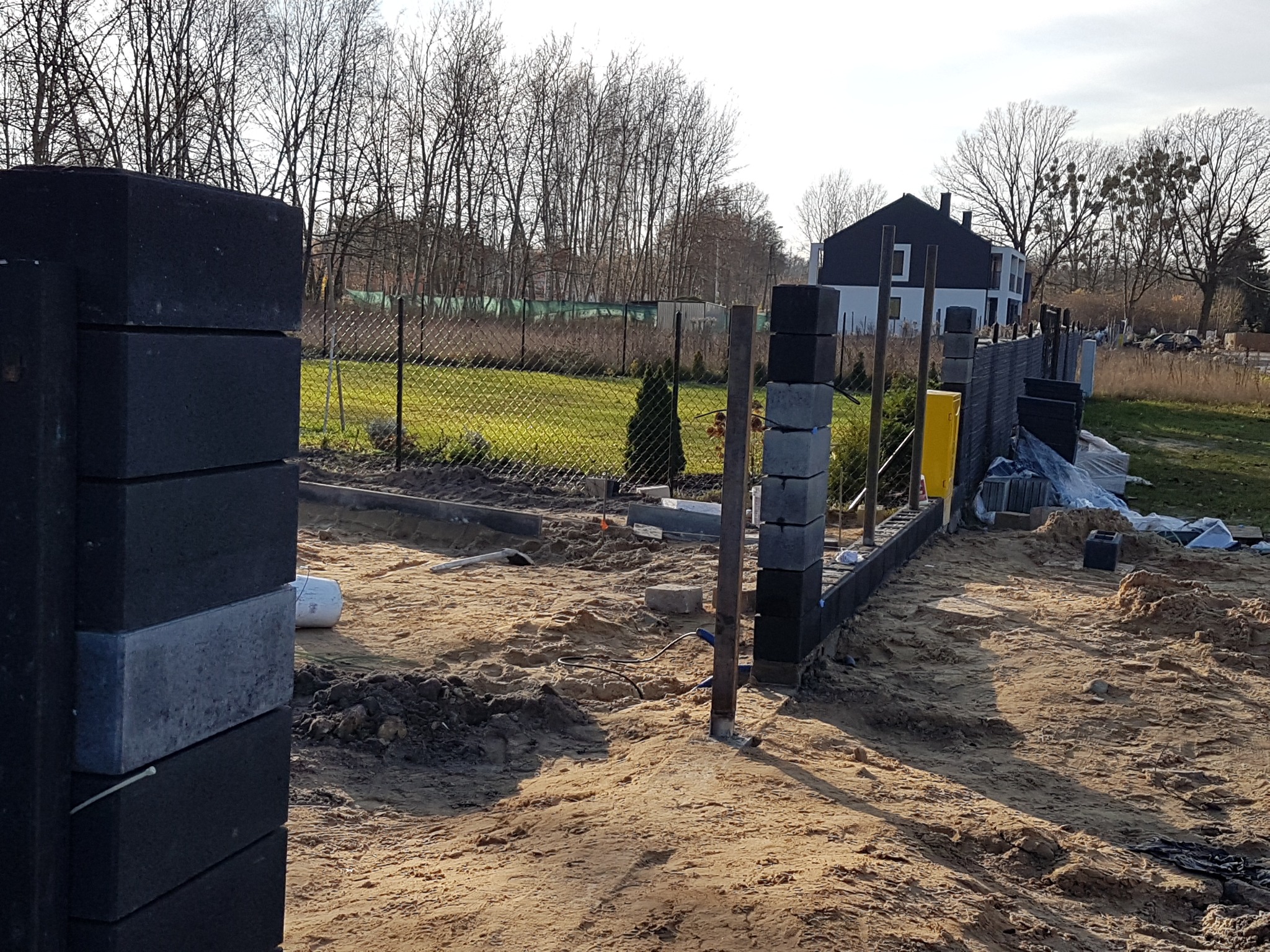 Budowa ogrodzenia z bloczków betonowych, widoczne słupki i podmurówka, teren budowy w trakcie prac ziemnych, w tle dom i drzewa.