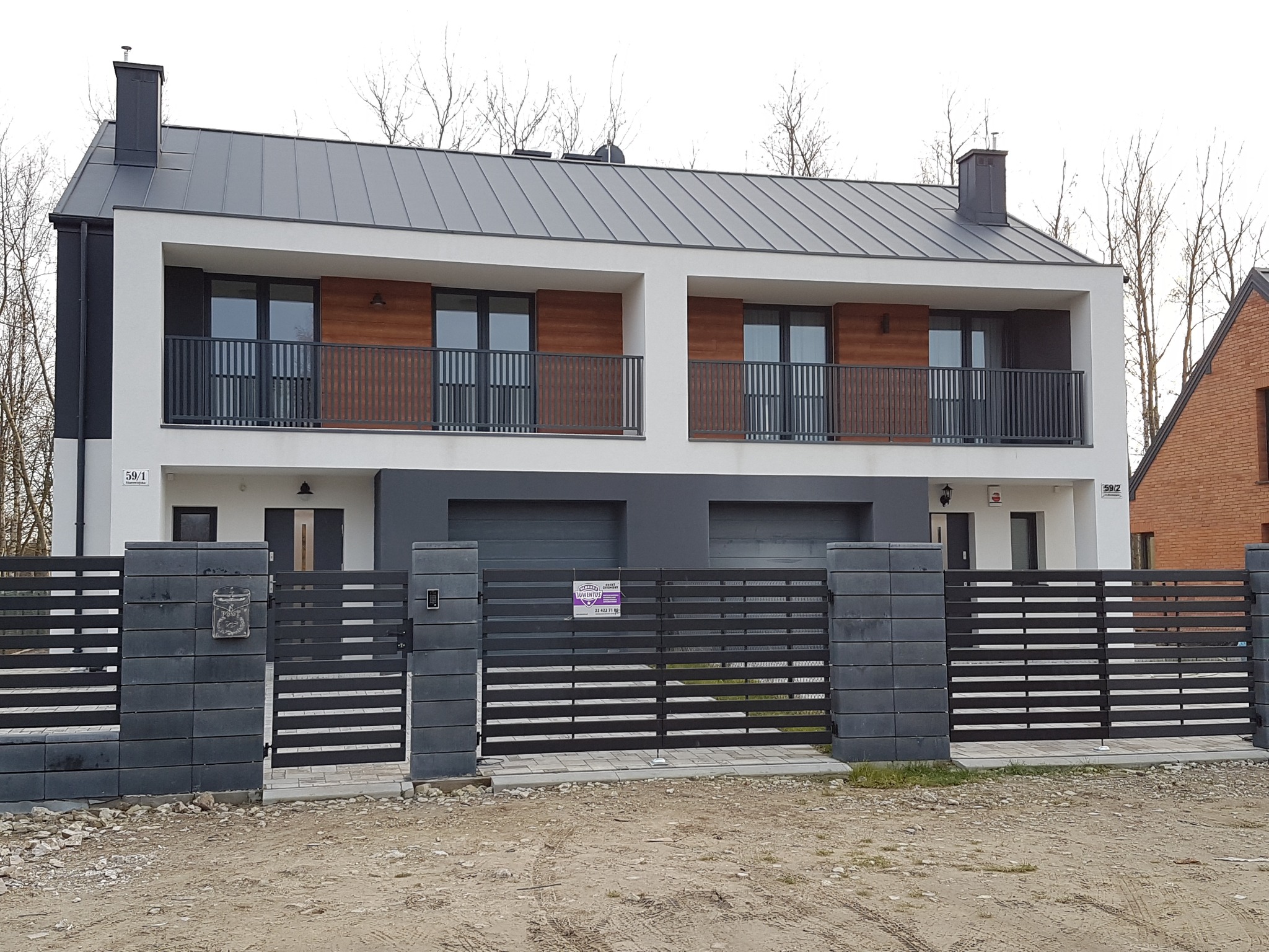 Nowoczesny bliźniak z szarą elewacją, drewnianymi panelami na balkonach i metalową bramą wjazdową z szarej kostki, widok z zewnątrz.