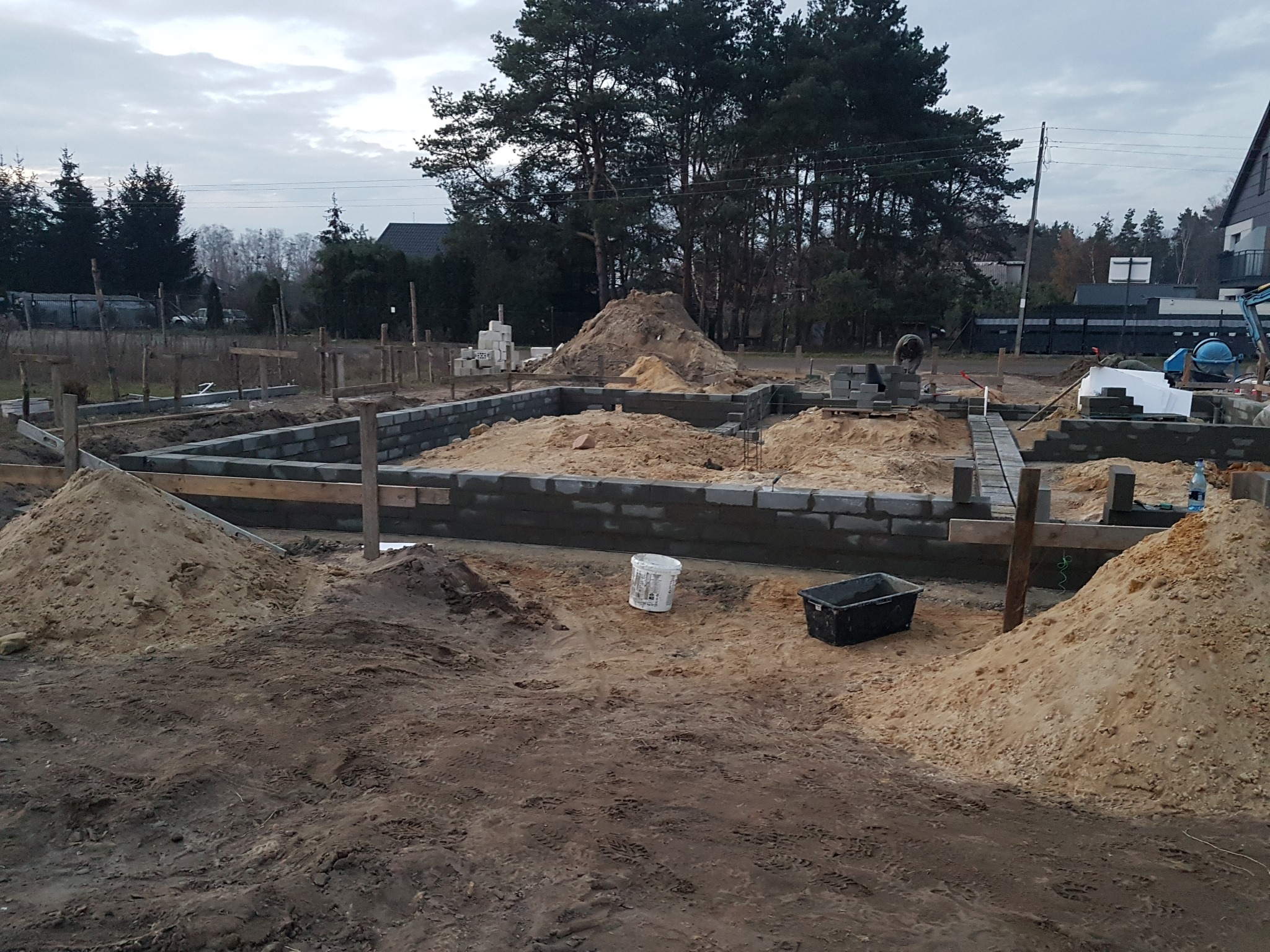 Budowa fundamentów domu jednorodzinnego: wzniesione ściany z bloczków betonowych, szalunki drewniane, piasek, narzędzia budowlane, pochmurne niebo.