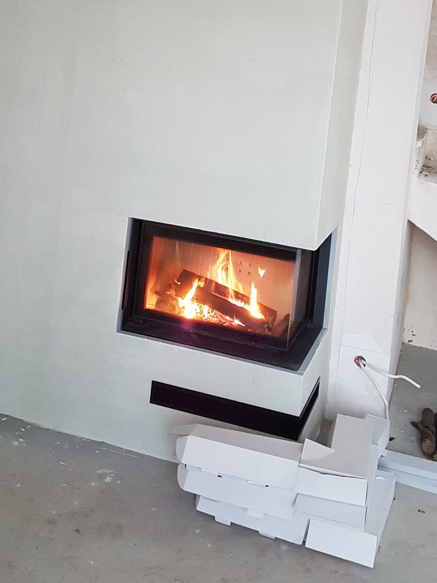 Nowoczesny kominek narożny z palącym się drewnem, obudowany jasnym, gładkim tynkiem. Na podłodze leżą białe elementy wykończeniowe oraz widoczne są ślady po pracach budowlanych.