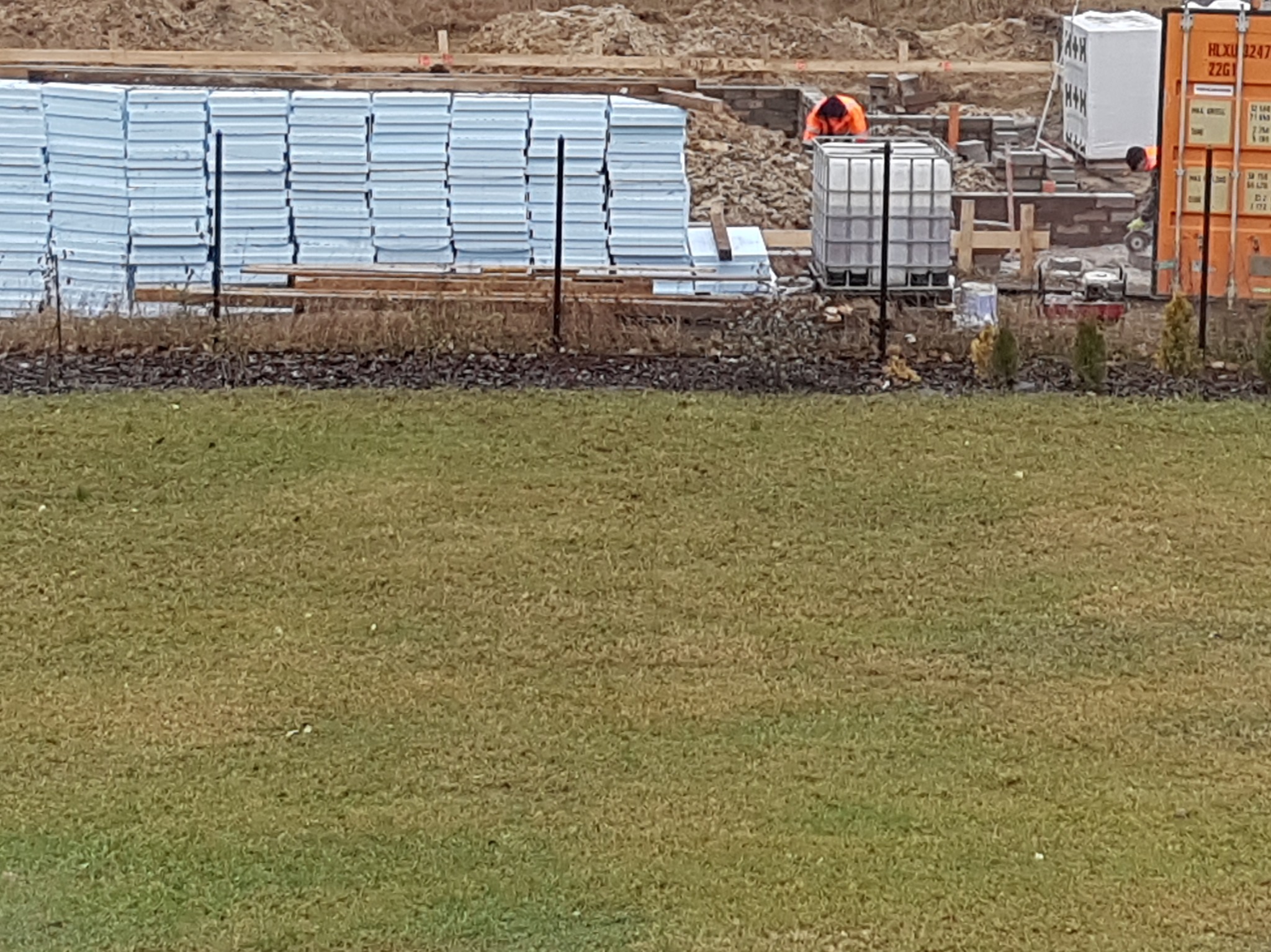 Styropianowe płyty izolacyjne ułożone na placu budowy, w tle pracujący robotnik w pomarańczowej kamizelce, kontener i zbiornik IBC.