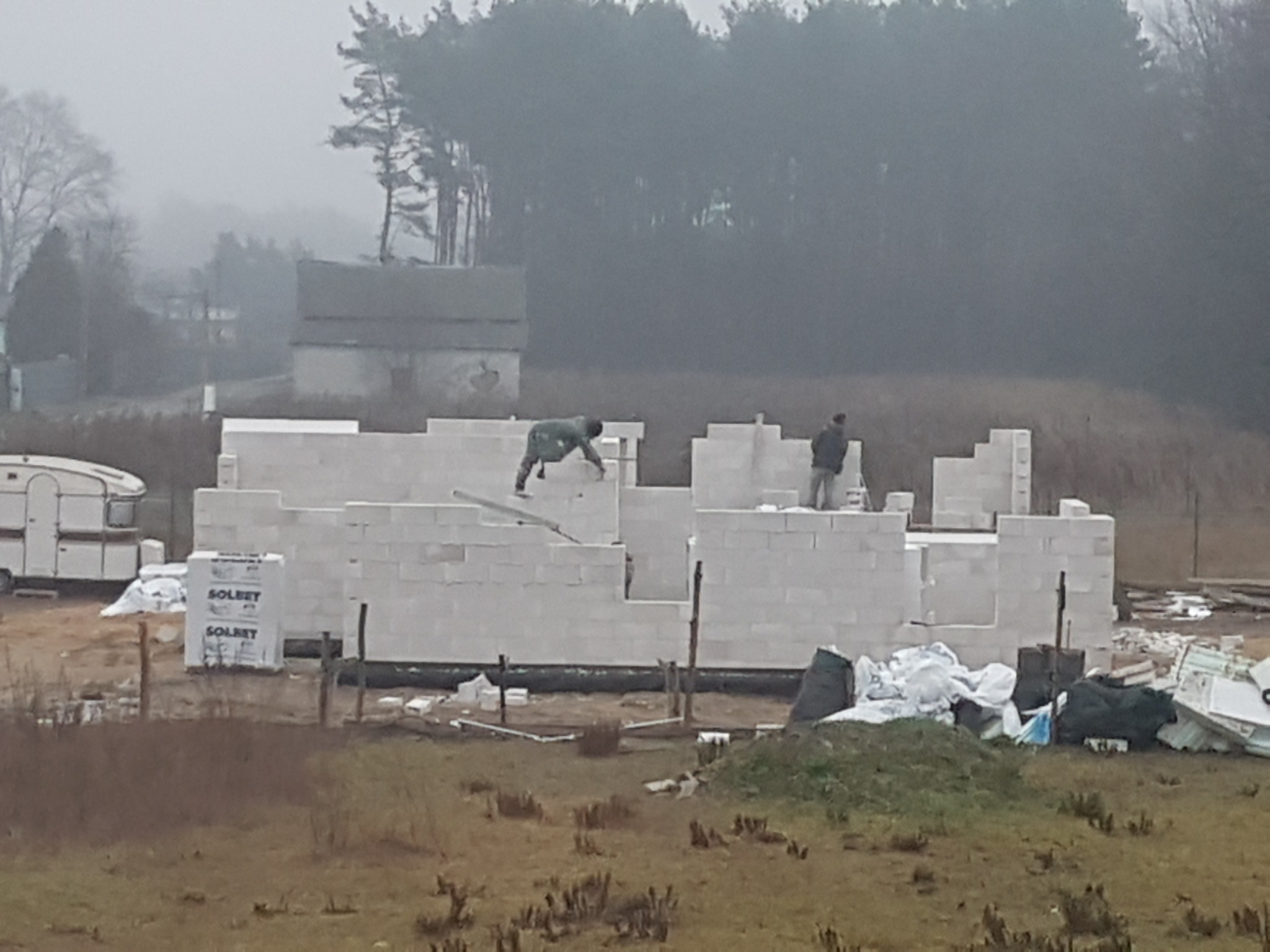 Wznoszenie ścian parterowego domu z białych bloczków betonowych, dwóch robotników w trakcie murowania, widok na działkę z przyczepą kempingową w tle, mglista pogoda.