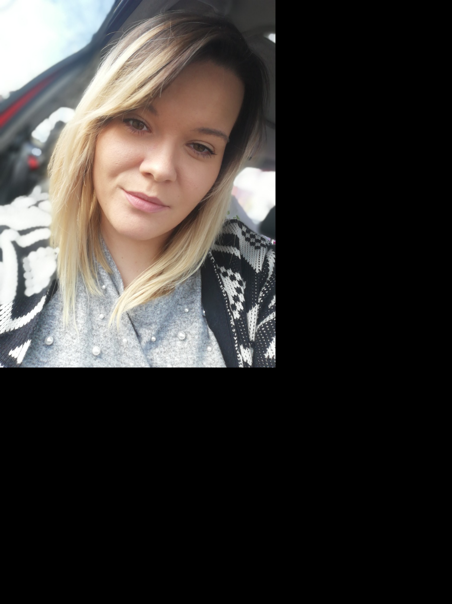 Portret kobiety z blond włosami w samochodzie, ubrana w szary sweter z perłami i czarno-biały kardigan.