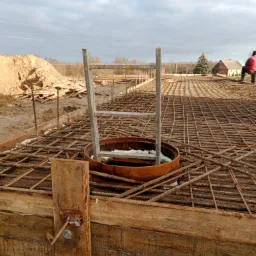 Zbrojenie płyty fundamentowej z drewnianym szalunkiem, widoczny właz kanalizacyjny i drabinka, w tle hałda ziemi i budynek mieszkalny.