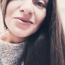 Portret kobiety z długimi brązowymi włosami, w szarym swetrze, z delikatnym uśmiechem, uchwycony z bliska.