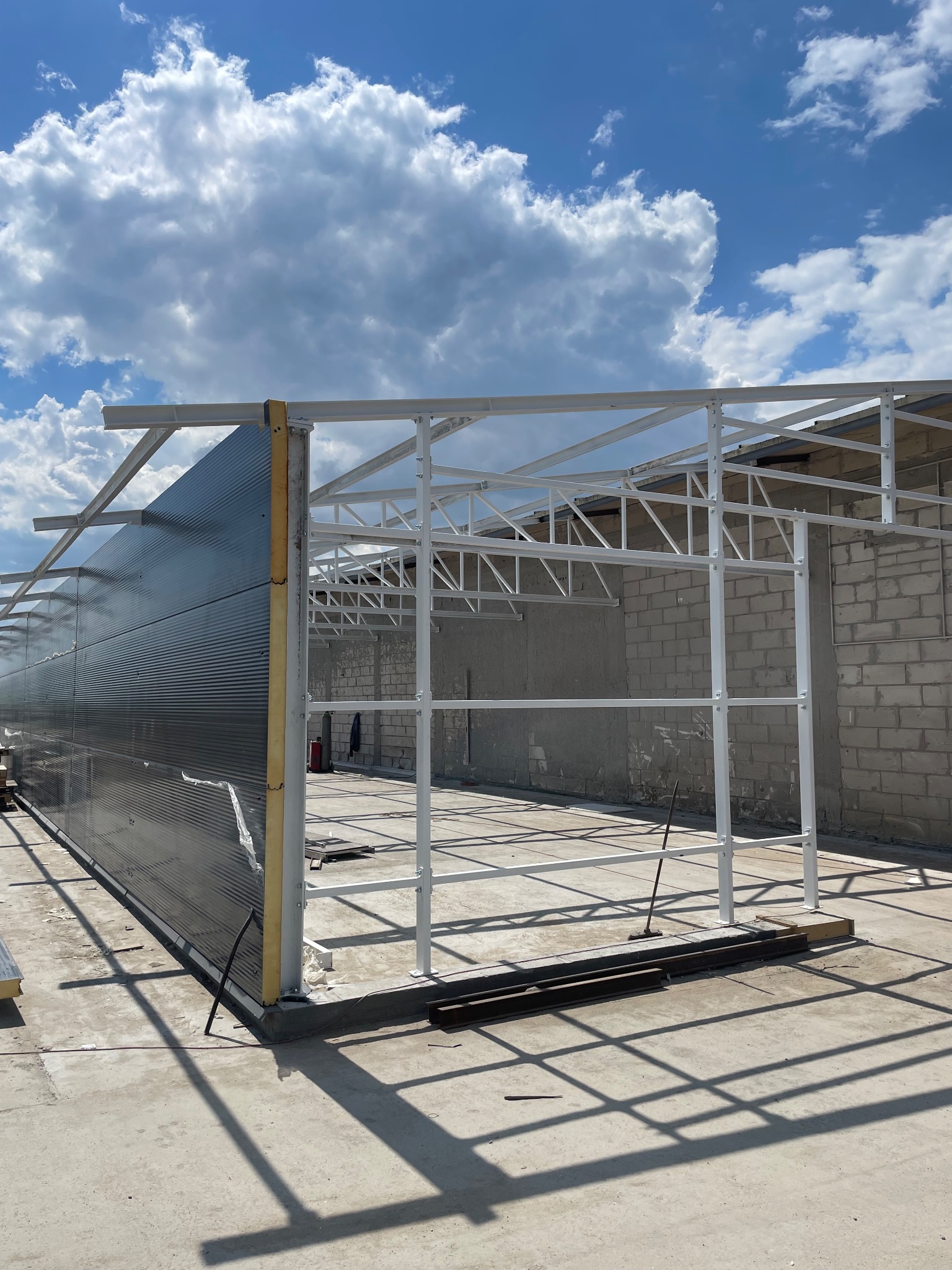 Konstrukcja hali w trakcie budowy: stalowy szkielet z zamontowanymi płytami warstwowymi, widoczne elementy mocujące. Słoneczny dzień, błękitne niebo z chmurami.