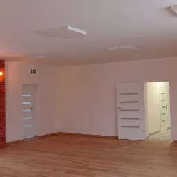 Puste, wykończone wnętrze z drewnianą podłogą, białymi ścianami, kwadratowymi panelami LED na suficie i dekoracyjną kolumną z czerwonej cegły z oświetleniem punktowym.