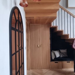 Katarzyna Kuczyńska Projektowanie Wnętrz - Klatka schodowa z drewnianymi schodami, zabudową pod schodami z siedziskiem i poduszkami, drewnianą podłogą ułożoną w jodełkę oraz lampami w kolorze miedzi.