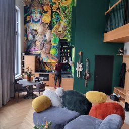 Katarzyna Kuczyńska Projektowanie Wnętrz - Wysokie, przestronne wnętrze z muralem Buddy, modułową sofą w kształcie kamieni, gitarami na ścianie i osobą przy blacie kuchennym.