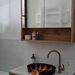 Katarzyna Kuczyńska Projektowanie Wnętrz - Umywalka nablatowa w kolorze granatowym z motywem słońca, bateria w kolorze mosiądzu, szafka pod umywalkę z drewnianymi frontami, lustro w drewnianej ramie z półką, oświetlenie punktowe w stylu retro.