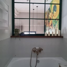 Katarzyna Kuczyńska Projektowanie Wnętrz - Biała wanna z ciemnobrązowym obramowaniem, bateria w stylu retro i dekoracyjne świece na parapecie okna z zieloną ramą, widok na sufit z okrągłymi otworami i fragment kolorowej grafiki na ścianie.