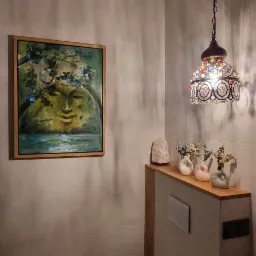 Umeblowana łazienka z toaletą, umywalką z czarnym blatem, obrazem z motywem twarzy i dekoracyjną lampą wiszącą z kolorowymi elementami.