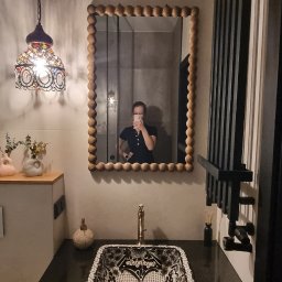 Katarzyna Kuczyńska Projektowanie Wnętrz - Umywalka nablatowa w czarno-białe wzory, rama lustra z drewnianymi kulkami, ozdobna lampa wisząca i grzejnik drabinkowy w łazience z beżowymi ścianami.