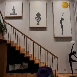 Salon z drewnianymi schodami, pod którymi znajduje się wnęka z siedziskiem i poduszkami, na ścianie wiszą trzy obrazy z motywami sylwetek i symboli słońca i księżyca, na podłodze parkiet w jodełkę.