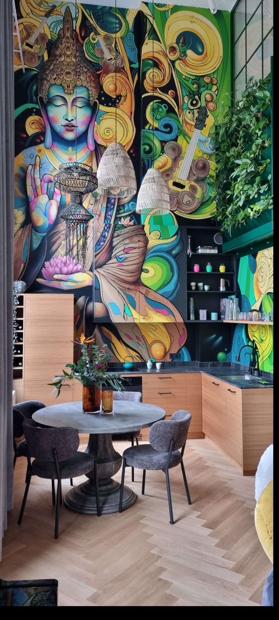 Jadalnia w Sopocie z okrągłym stołem i krzesłami na parkiecie w jodełkę, na ścianie mural z motywem Buddy i abstrakcyjnymi wzorami, rattanowe lampy wiszące, drewniane szafki kuchenne i półki...