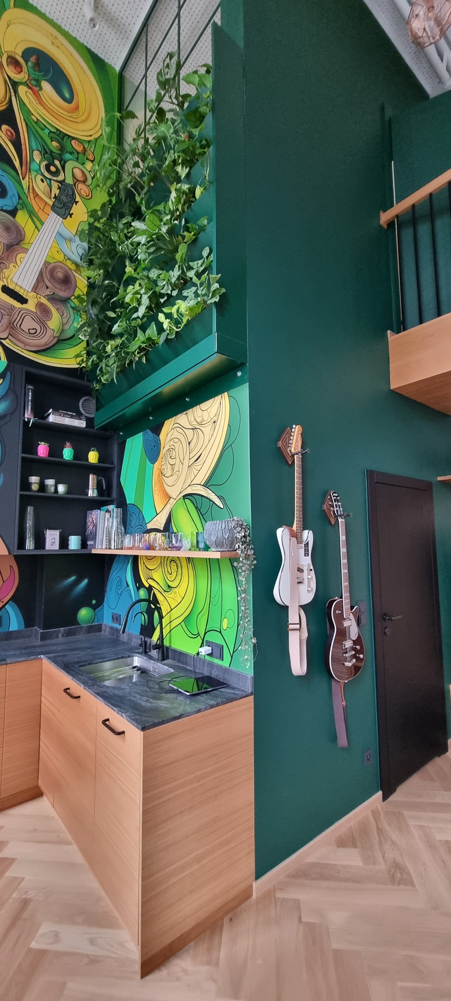 Nowoczesna kuchnia z drewnianymi szafkami, czarnym blatem i kolorowym muralem na ścianie, uzupełniona o wiszące gitary.