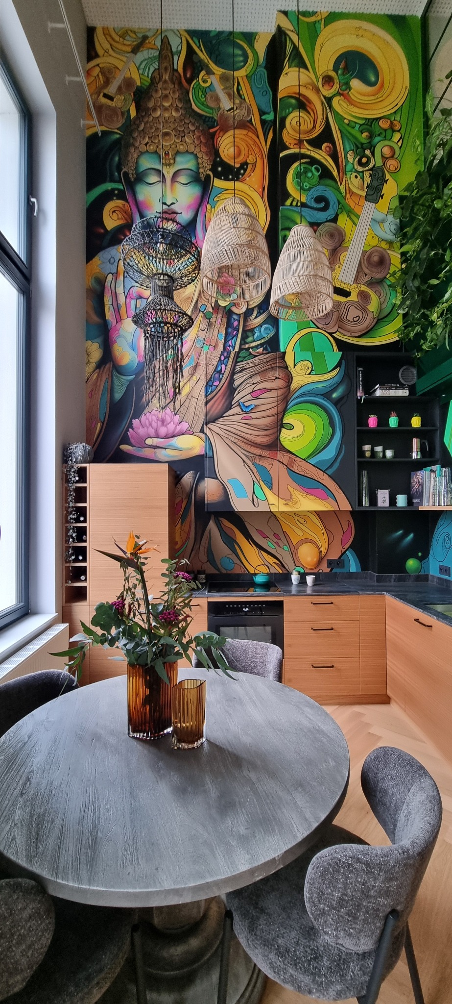 Jadalnia z okrągłym stołem, szarymi krzesłami i muralem z wizerunkiem Buddy w tle, uzupełniona drewnianą zabudową kuchenną i lampami z wikliny.