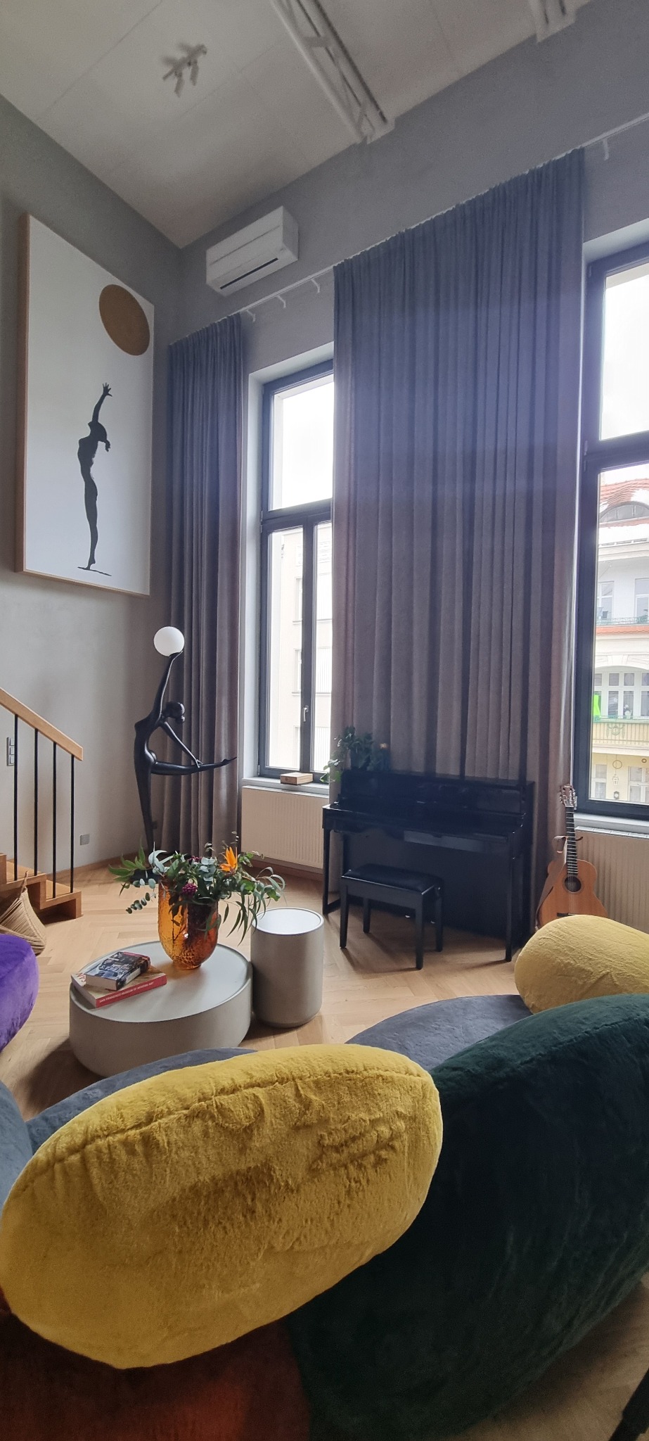 Wysokie pomieszczenie z dużymi oknami zasłoniętymi szarymi zasłonami, pianino, gitara, okrągły stolik kawowy z wazonem kwiatów i rzeźbą baletnicy z lampą, obraz z motywem tanecznym na ścianie.