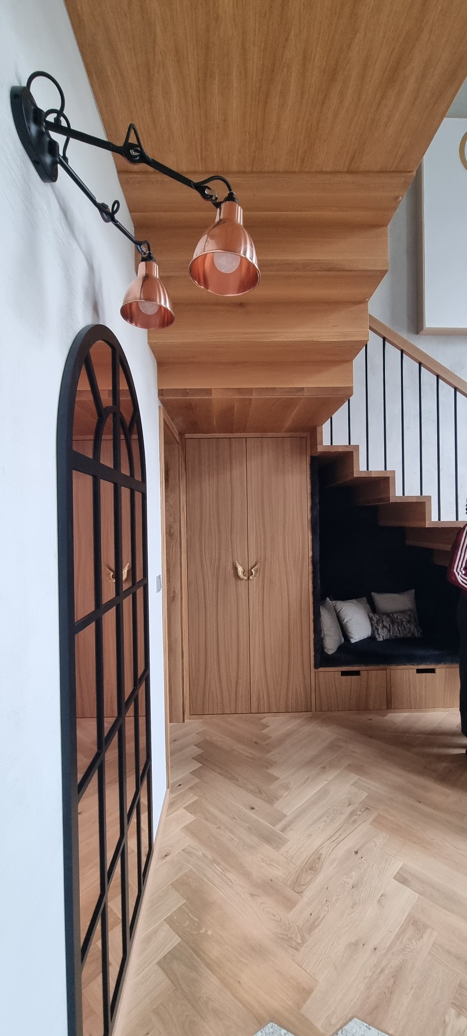 Klatka schodowa z drewnianymi schodami, zabudową pod schodami z siedziskiem i poduszkami, drewnianą podłogą ułożoną w jodełkę oraz lampami w kolorze miedzi.