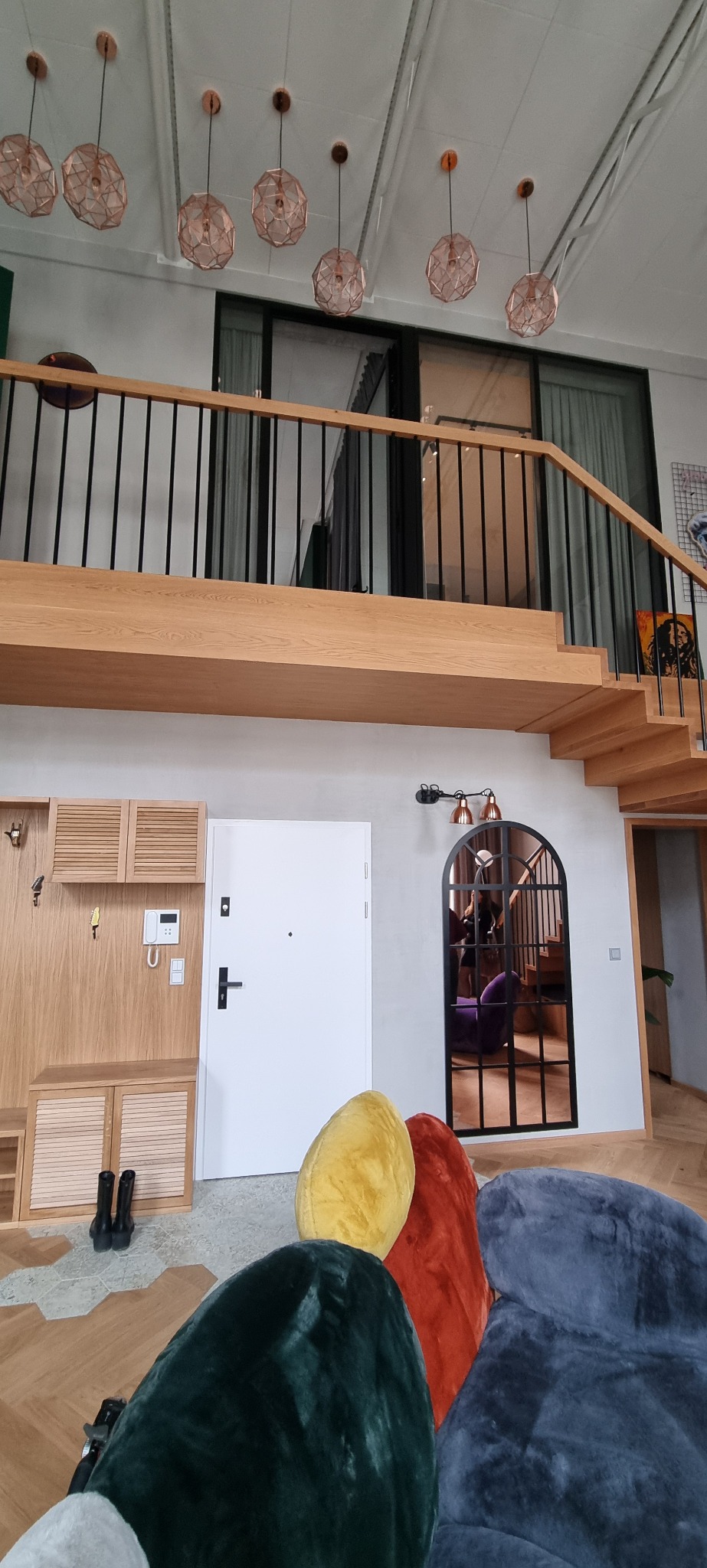 Wysokie wnętrze z antresolą, drewnianymi schodami, zabudową szafek w przedpokoju, białymi drzwiami wejściowymi i dekoracyjnymi, kolorowymi pufami w kształcie kamieni.