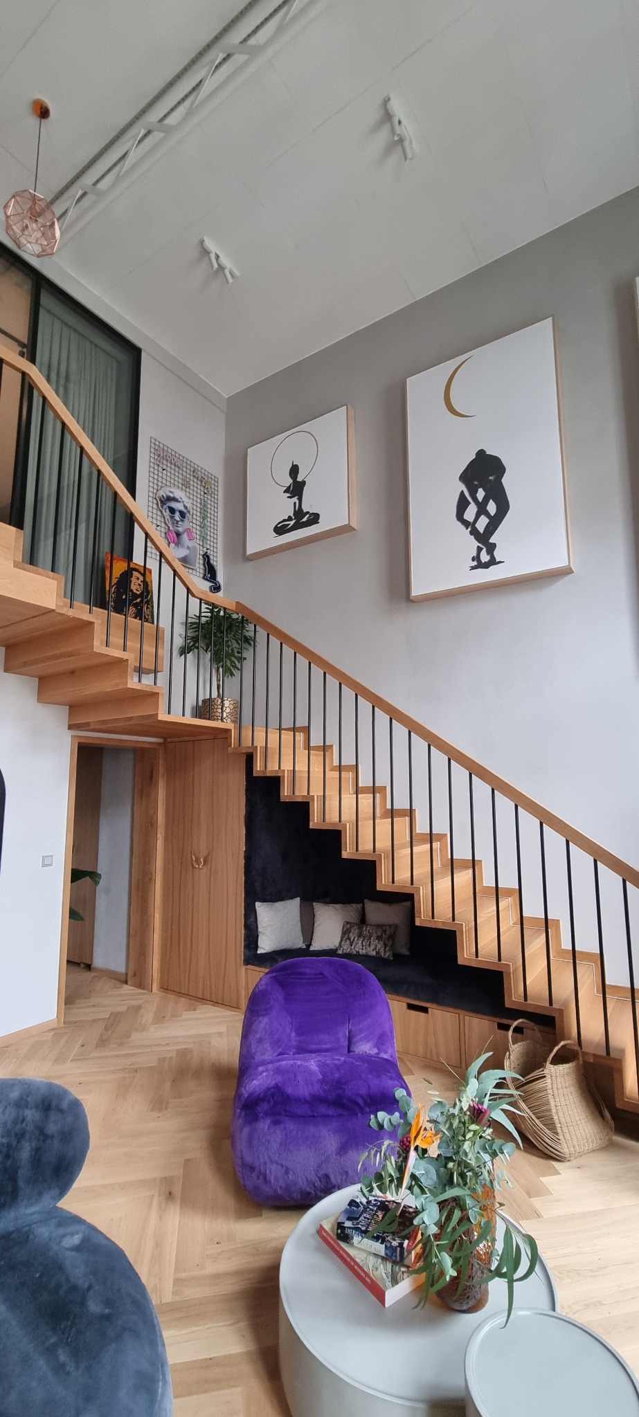 Dwupoziomowe wnętrze z drewnianymi schodami z czarną balustradą, wbudowanym siedziskiem z poduszkami pod schodami, fioletowym pufem i nowoczesnymi obrazami na ścianie. Na podłodze parkiet ułożony...