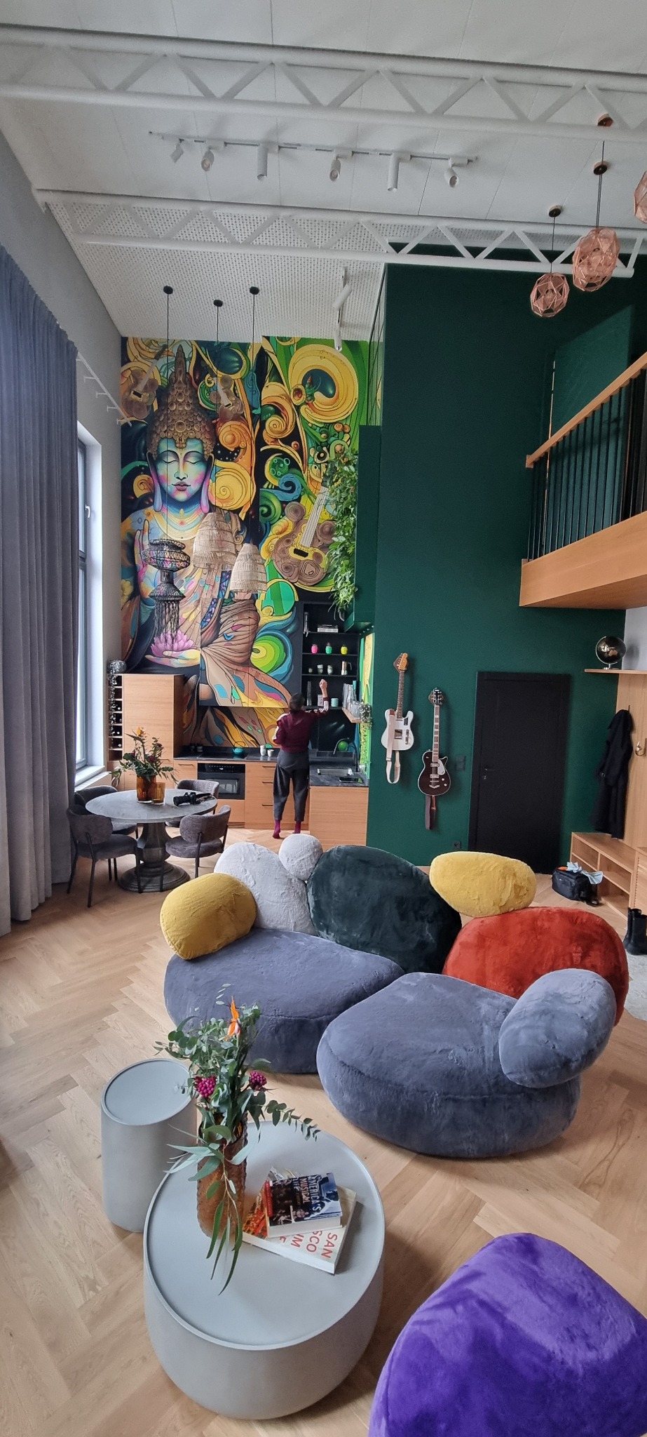 Wysokie, przestronne wnętrze z muralem Buddy, modułową sofą w kształcie kamieni, gitarami na ścianie i osobą przy blacie kuchennym.