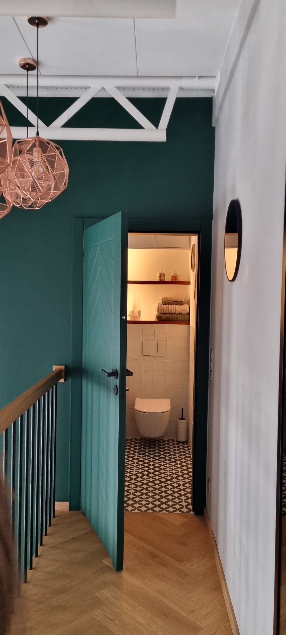 Otwarta, turkusowa łazienka z wzorzystą podłogą, widziana z korytarza z drewnianą podłogą ułożoną w jodełkę, ciemnozielone ściany i ozdobne lampy.